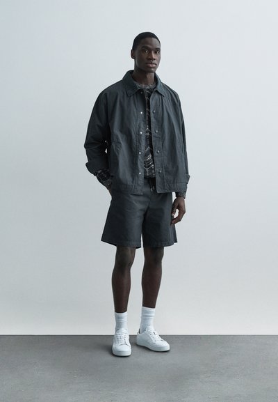 Ciemnoszara kurtka z zatrzaskami, wzorzysta koszula, pasujące szorty, białe trampki i skarpety za kostkę. Gładka tkanina o casualowym kroju.