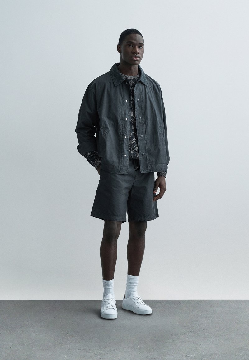 Dark gray jacket with snap buttons, patterned shirt, matching shorts, white sneakers, and crew socks. Smooth fabric with a casual design.