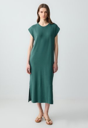 Türkisgrünes, knielanges Kleid aus weichem Stoff mit kurzen Ärmeln und einem seitlichen Schlitz. Getragen mit braunen flachen Sandalen.