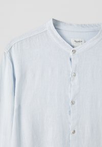 Jasnoniebieska lniana koszula z okrągłym dekoltem, zapinana na guziki, o teksturowanym wykończeniu. Prosty fason bez wzorów i ozdób.