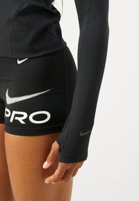 Melnas garās piedurknes krekls un kompresijas šorti. Krekls ar pieguļošu dizainu un gludu tekstūru; šortiem ir balts "Nike Pro" zīmols un logo.