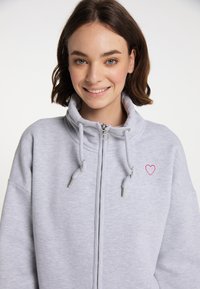 Šedá mikina na zip má vysoký límec, šňůrky, kovové detaily a malou červenou srdíčko vyšitou na levé straně.
