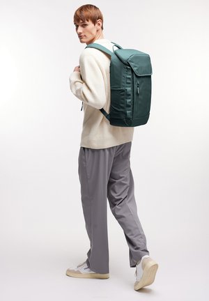 Zaino verde con una forma strutturata, realizzato in materiale liscio, dotato di chiusura a pattina, tasca anteriore con zip e spallacci imbottiti.