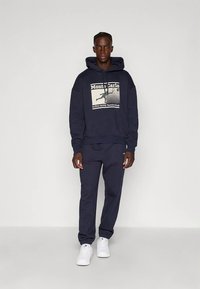 Uomo che indossa un hoodie blu navy con una grafica di tennis "Monte Carlo", pantaloni della tuta abbinati e sneakers bianche, in piedi di fronte a uno sfondo bianco uniforme.