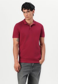 Mand iført en bordeaux polo skjorte og grå jeans står med den ene hånd i lommen mod en hvid baggrund.