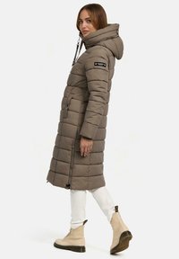 Lange, pufferjas in gedempt bruin met een capuchon, voorzien van horizontale stiknaden, een ritssluiting en twee voorkruiszakken, gecombineerd met lichtgekleurde laarzen.