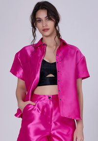 Chemise en satin rose à manches courtes avec un col et des boutons noirs, portée par-dessus un top noir court, associée à un pantalon en satin rose assorti.