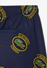 Pantalones cortos navy con cinturilla elástica que presentan parches del logo de Lacoste en verde, negro y amarillo, con un detalle de bolsillo trasero.
