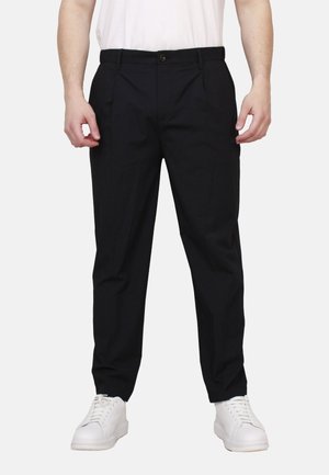 Uomo che indossa pantaloni neri a gamba dritta, sneakers bianche e una t-shirt bianca, in piedi davanti a uno sfondo chiaro e semplice.