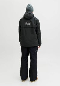 Isik mustas TECH FUSION jakkis, mustades pükstes, halli mütsiga ja helepruunide kingadega, seisab seljaga kaamera poole valge tausta ees.