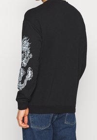Černá mikina s dlouhými rukávy, na levém rukávu zdobená detailním bílým potiskem draka. Vyrobena z měkké, texturované látky.