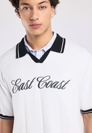 Jonge man die een witte korte mouwtrui draagt met een zwarte kraag en "East Coast" geborduurd op de voorkant over een gestreept overhemd.