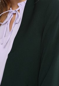 Zelený blejzr s hladkým povrchem a otevřeným předním dílem, který je spárován se světle levandulovou blůzou s volánovým výstřihem a tými.