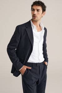 Donkerblauwe blazer met een gestructureerde afwerking, enkele knoopsluiting, gecombineerd met een wit overhemd met kraag en op maat gemaakte broek.