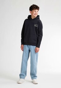 Černá mikina s kapucí a přední kapsou, s malým bílým grafickým motivem. Kombinována se světlými volnými džínami a bílými teniskami.