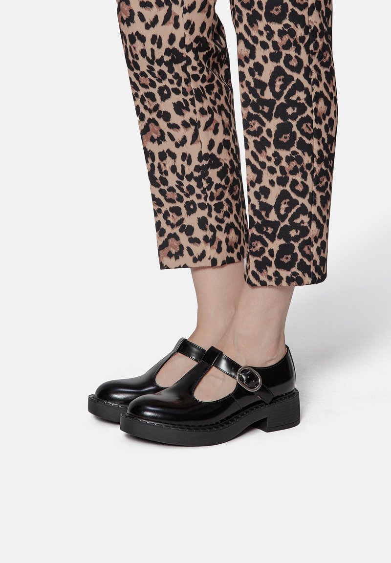 Des chaussures T-bar en cuir verni noir avec une semelle épaisse, un détail de boucle circulaire et une finition brillante, associées à un pantalon court à imprimé animal.