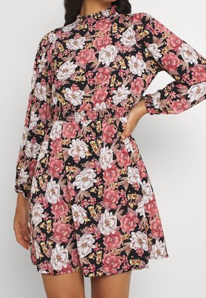 Femme portant une robe longue noire à manches longues avec des fleurs roses, blanches et beiges et une taille élastique, posant avec une main sur la hanche.