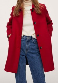 Femme portant un manteau rouge à boutons sur un pull à col roulé blanc, les mains dans les poches et un jean bleu taille haute, sur fond uni.