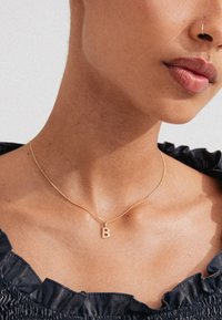 Goldkette mit einem kleinen Pendant in Form des Buchstabens "B", getragen auf einem nackten Hals, ergänzt durch ein gerafftes Oberteil aus dunklem Stoff.