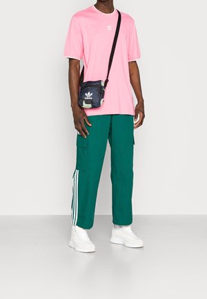 Mężczyzna w różowej koszulce Adidas, zielonych spodniach dresowych Adidas z białymi paskami, białych trampkach i czarnej torbie na ramię Adidas.