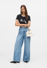 T-shirt nera con dettagli bianchi e stampa grafica, jeans leggeri blu chiaro larghi, ballerine nere con borchie e una piccola borsa color crema con logo.