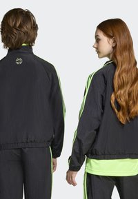 Crne vjetrovke s neonsko zelenim detaljima i prugama, teksturiranog završetka i elastičnim manžetama, nošene preko uparene hlače.