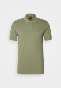 Polo manches courtes vert olive avec patte à deux boutons et petit écusson carré sur la poitrine gauche sur fond blanc.