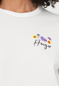 Λευκό βαμβακερό μπλουζάκι με λουλουδάτο σχέδιο με κίτρινα και μωβ λ�ουλούδια και το όνομα της μάρκας "Hugo" σε μαύρη γραφή στο αριστερό στήθος.