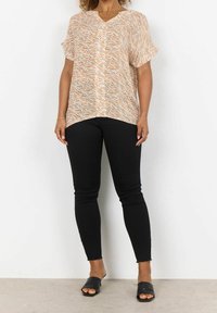 Blusa a maniche corte con scollatura a V di colore arancione chiaro con un motivo ondulato, abbinata a jeans neri aderenti e sandali neri scivolati.