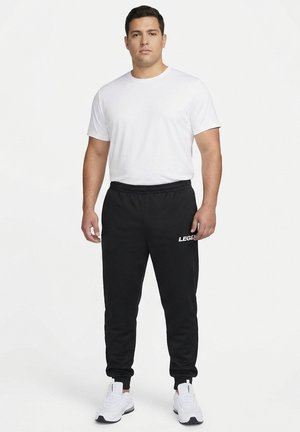 Man standing wearing a plain white t-shirt, black joggers with "LEGEND" text, and white sneakers against a plain white background.