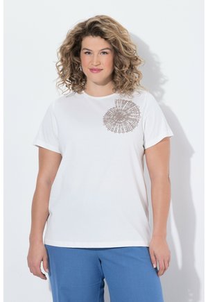 Kvinde med krøllet hår iført hvid kortærmet skjorte med cirkulært blonderdetalje på brystet og blå bukser, stående mod en hvid baggrund.