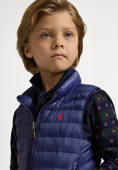 Marineblå vattert vest med horisontal quiltet tekstur, glidelåslukking og rødt brodert logo. Under er en mønstret langermet skjorte med små motiver.