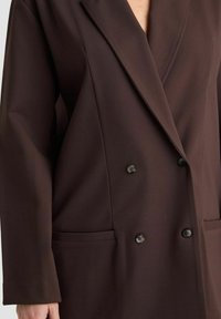 Dunkelbrauner doppelt-geknöpfter Blazer mit Reverskragen, vier Knöpfen und aufgesetzten Vorderaschen auf einer Person.