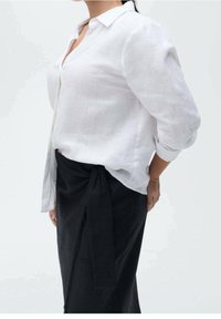 Chemise en lin blanc à manches longues, avec des boutons sur le devant et un col, associée à une jupe portefeuille noire avec un détail noué à la taille.