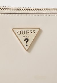 Materiał w kolorze kremowym o gładkiej fakturze, zdobiony trójkątnym logo GUESS w złotym i czarnym kolorze z motywem znaku zapytania. Widoczny detal zamka błyskawicznego.