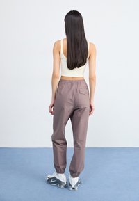 Žena s dugom kosom nosi bež crop top i svijetlosmeđe hlače, okrenuta leđima na plavom podu uz običan bijeli zid.