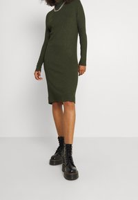 Abito lungo a maniche lunghe in verde oliva con righe, collo alto, silhouette aderente e orlo sino al ginocchio, abbinato a stivali da combattimento neri stringati.