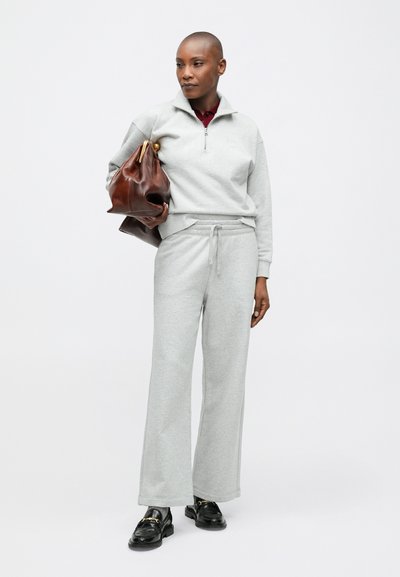 Sivý svetrier s polovičným zipsom a zúženými manžetami, doplnený o zodpovedajúce tepláky. Modelka drží hnedú koženú kabelku a má na sebe čierne mokasíny.