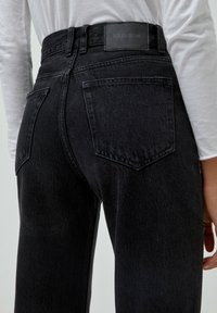 Jean en denim noir avec une taille haute, des poches arrière et une étiquette de marque sur la ceinture. Le tissu a une texture lisse.
