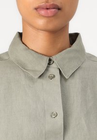 Olive green linen shirt featuring a pointed collar, button-down front with two buttons, and a textured fabric finish.