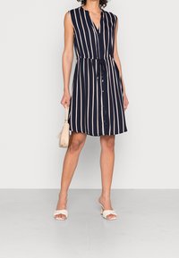 Vestido de marina con rayas verticales en color crema, diseño sin mangas, con botones en el frente y cintura ajustable con cordón. Acompañado de un bolso ligero y sandalias.