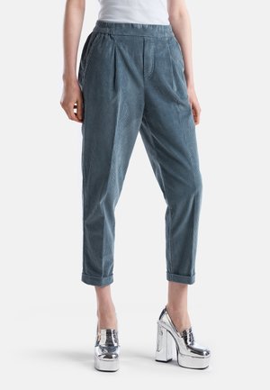 Personne portant un pantalon court en velours côtelé bleu-gris et des chaussures à plateforme argentées métalliques à talons hauts, debout sur fond blanc.