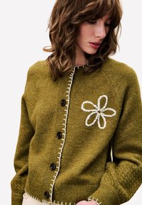 Olijfgroene gebreide cardigan met een bloemige witte borduursel, voorzien van een knoopsluiting aan de voorkant, geribbelde mouwen en contrasterende witte bies langs de knopen.