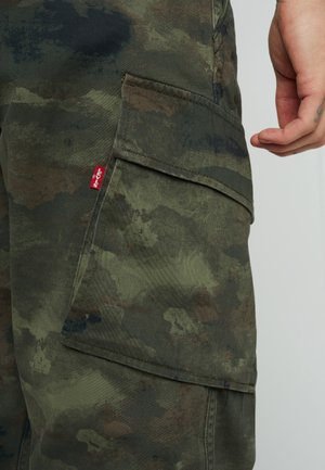Nærbillede af en hånd ved en grøn og brun camouflage cargo bukselomme med et lille rødt Levi's mærke synligt på lommekanten.