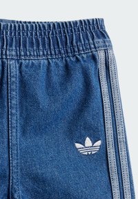 Džinsiniai džogai su elastinga juosmena, turintys baltas trijų juostelių detales ir mažą baltą logotipą kairėje šlaunies pusėje.