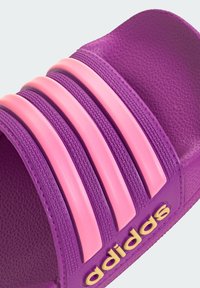 Fioletowa klapka na ślizgu z trzema różowymi paskami i fakturowaną powierzchnią. Logo w złotym kolorze z boku, z szerokim paskiem.