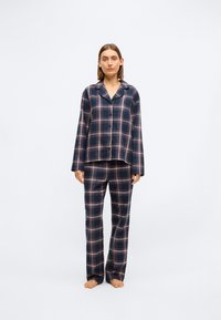 Navy sinisessä ruutukuviossa oleva pyjama-setti, joka sisältää pitkähihaisen kauluksellisen paidan ja siihen sopivat housut. Pehmeää kangasta, jossa on valkoisia ja punaisia viivoja ruutukuviolla.
