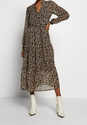 Abito lungo a maniche lunghe in chiffon nero con orlo a strati, caratterizzato da una scollatura a V e motivi floreali beige. Indossato con stivali alla caviglia bianchi.