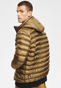 Brązowa kurtka puchowa z kapturem, z poziomymi przeszyciami, zapinanymi na zamek kieszeniami bocznymi oraz naszywką z logo na dolnej lewej stronie.