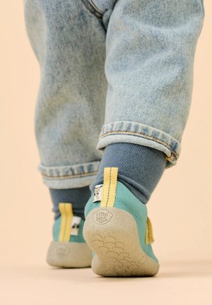 Kleinkind trägt blaue Socken, aufgerollte helle Denim-Jeans und türkisfarbene Schuhe mit gelben Zuglaschen, steht auf einem beigen Boden.